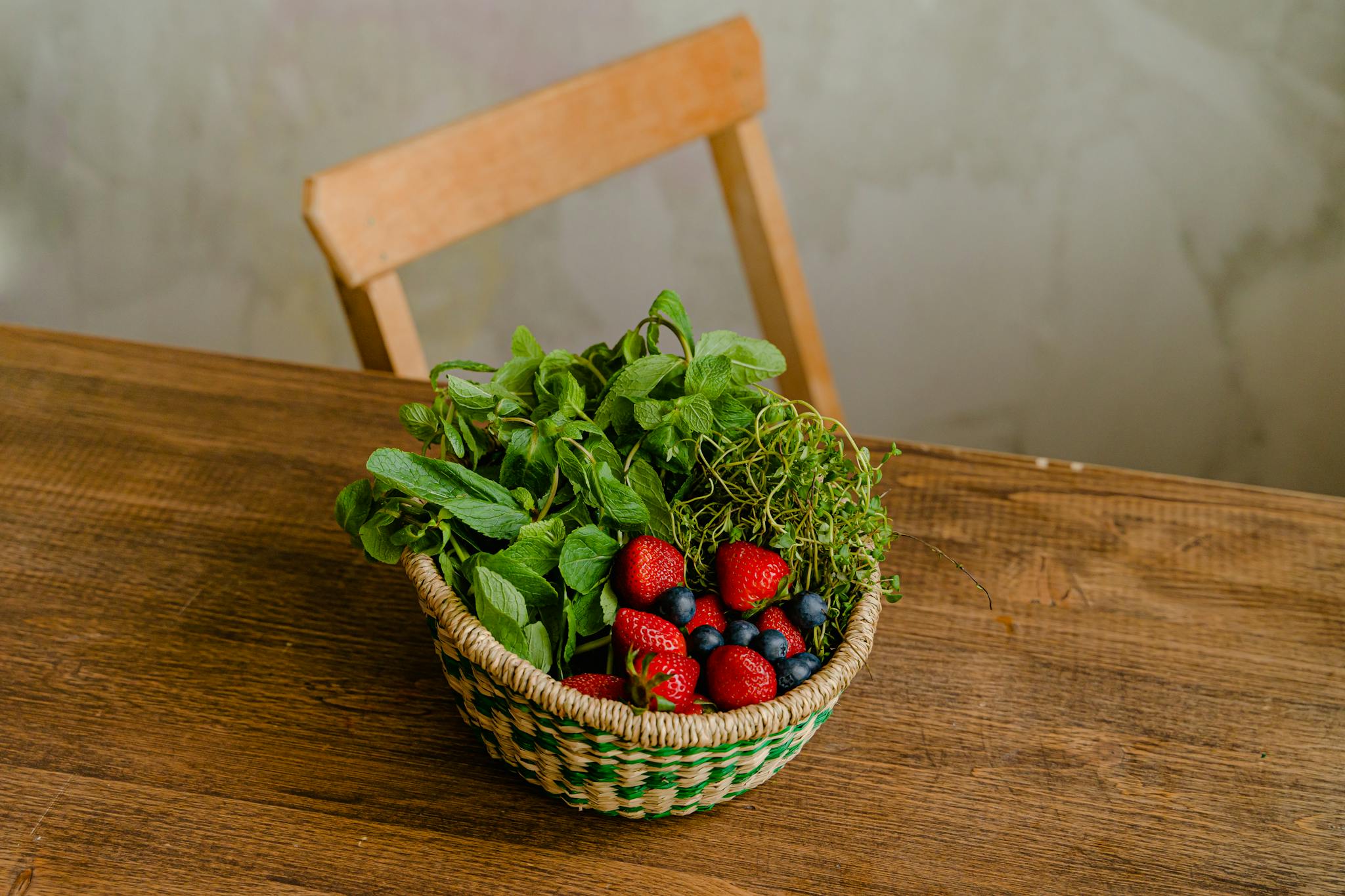 A woven basket filled with mint, strawberries, and blueberries on a wooden table.