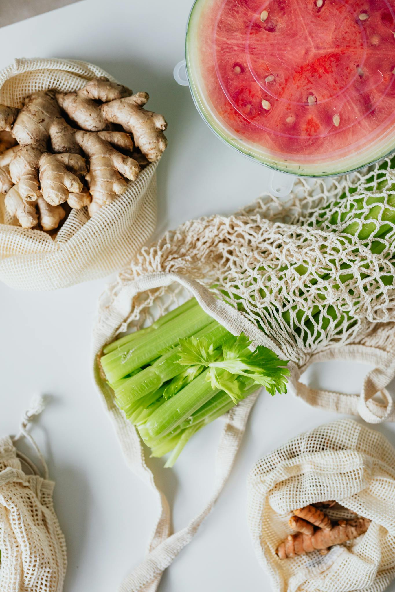 Top view of fresh ginger, watermelon, celery, and turmeric in mesh bags on a white counter.